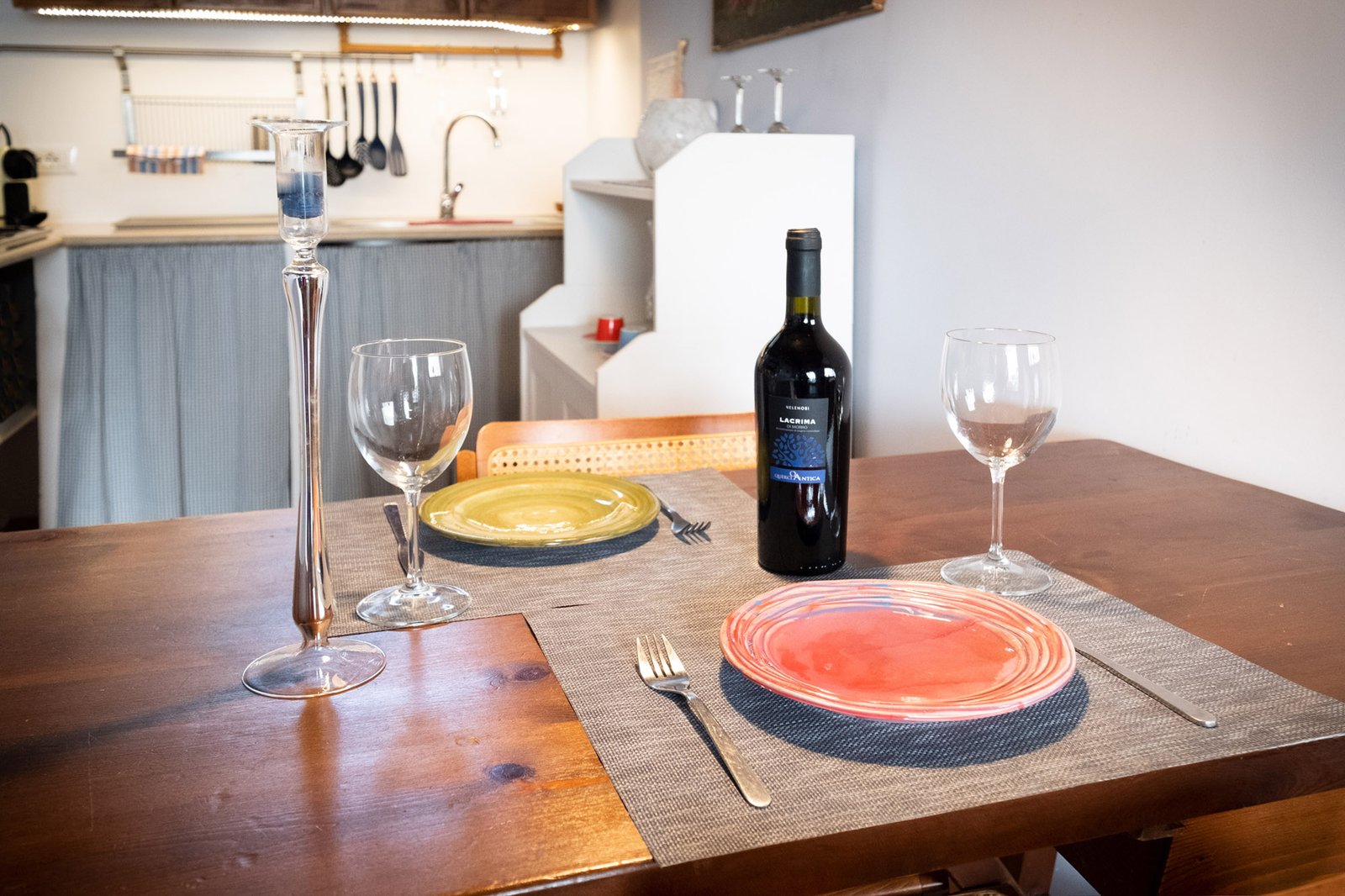 tavola della cucina apparecchiata per il pasto con piatti colorati, vino e candela, sullo sfondo il lavandino illuminato